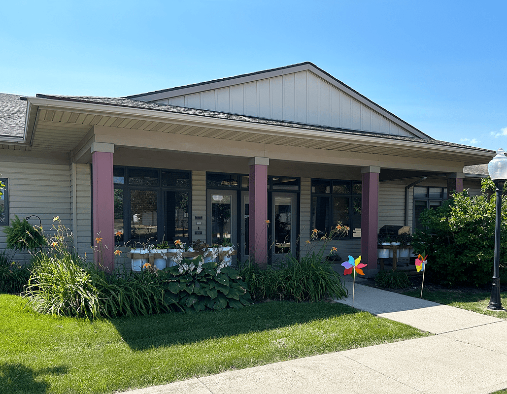 A single-story assisted living community building with a covered front porch supported by purple columns. There are flower beds with plants and flowers, colorful pinwheels planted in the ground, a well-maintained lawn, a sidewalk leading to the entrance, and a lamppost on the right side. The sky is blue with a few clouds.