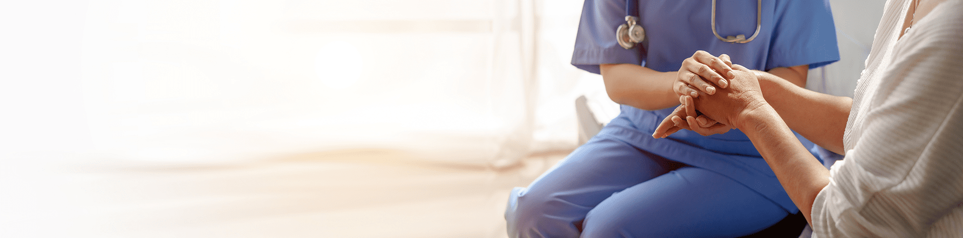 Close-up of a healthcare professional in blue scrubs holding and comforting an elderly patient's hand in a well-lit medical setting.