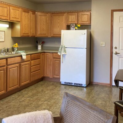 Kitchen with wooden cabinets, a white refrigerator with a cloth hanging on the handle, a countertop with a yellow flower and utensils, and a beige floor. There is a door and a chair on the right side.