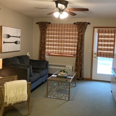 Living room with a black sofa, a glass coffee table, and a wooden chair with a yellow blanket. There are patterned curtains on the window and door, a ceiling fan with lights, and wall art featuring utensils.