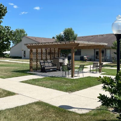 A wooden pergola with lattice sides providing shade over a seating area with chairs and a table, situated on a grassy lawn in a residential community. There are sidewalks, houses, and a lamppost nearby under a clear blue sky.