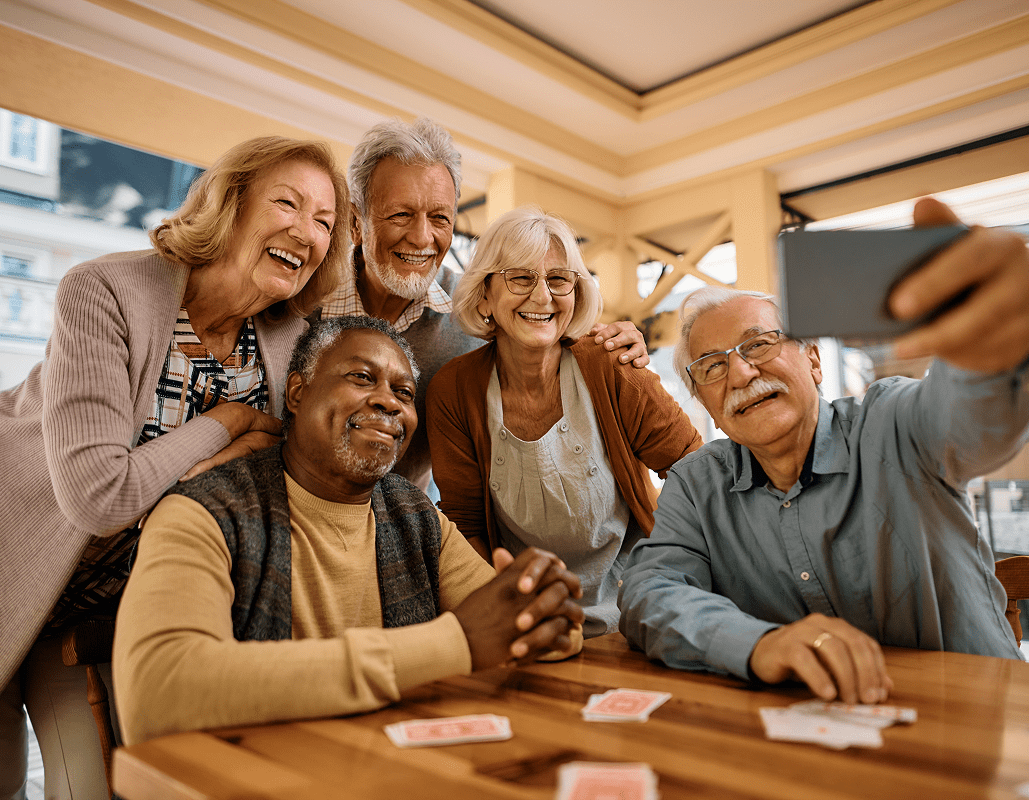 Six elderly people are gathered around a table, smiling and taking a selfie together in a well-lit indoor setting, possibly a community center or cafe.