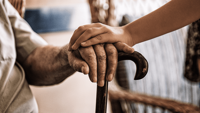 A young person’s hand gently holding an elderly person’s hand, which rests on a cane, symbolizing care and support in a caregiving setting.