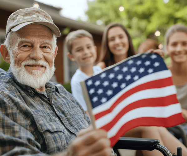 An elderly man with a white beard and gray cap smiling and holding an American flag, with three young people smiling in the background outdoors.