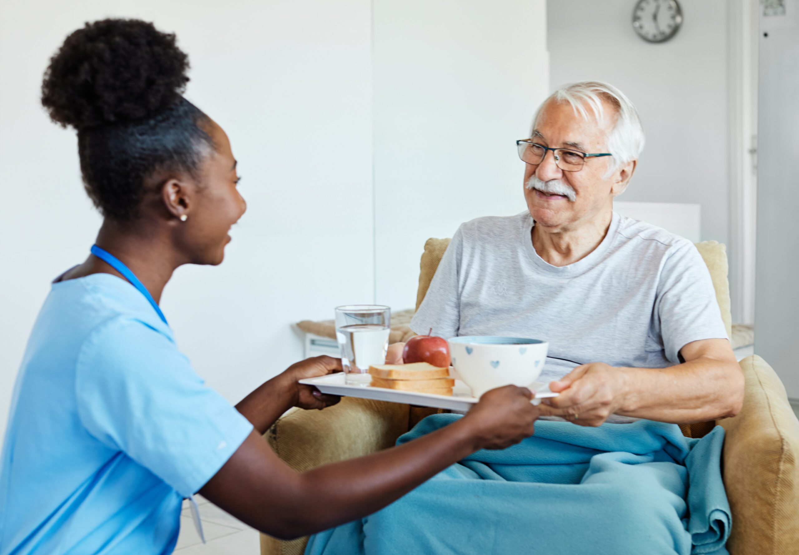 A caregiver in blue scrubs offers a tray with a bowl, apple, toast, and a glass of water to an elderly man sitting comfortably in a beige armchair, smiling.
