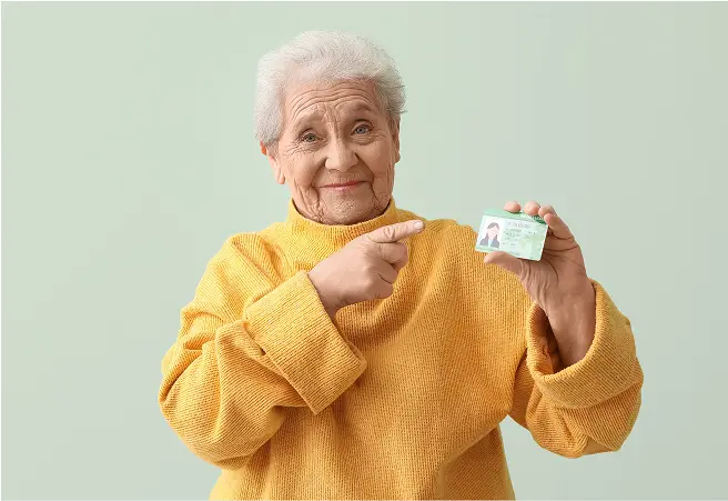 An elderly woman with white hair, wearing a yellow sweater, smiling and pointing at a driver's license she is holding in her right hand against a light green background.