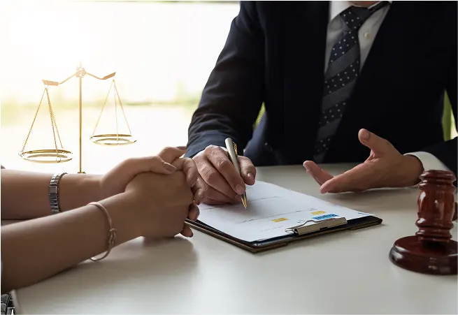 A person in a suit is sitting at a desk, signing a document with a gold pen, while another person’s hands are clasped in front of them. A gavel and a set of scales are visible in the background, suggesting a legal or official setting.