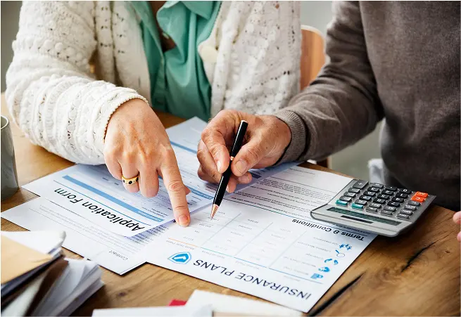 Two people are sitting at a table reviewing insurance paperwork; one person is pointing at a form while the other holds a pen, with a calculator nearby.