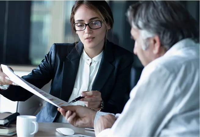 A young woman with glasses, dressed in a dark blazer and white shirt, is showing documents to an older man with gray hair, who is wearing a white shirt. They are seated at a table in a professional setting, discussing the documents.
