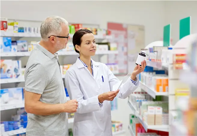A pharmacy technician and an older man are standing in a pharmacy aisle, with the technician holding a medication bottle and explaining it to the man. Shelves stocked with various medication boxes are visible in the background.